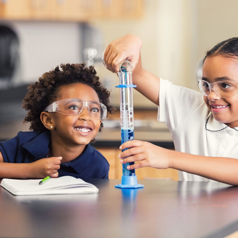 African American elementary school girl is smiling while sitting at desk in science lab. She is watching her classmate combine chemicals during science experiment. Little girls are students in private elementary school, and are wearing school uniforms. They are excited and having fun during class in modern science lab classroom.